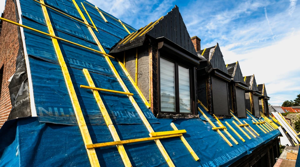 Photographie réaliste en extérieur montrant un couvreur expérimenté travaillant sur une toiture de maison résidentielle classique. L'artisan, vêtu de tenue professionnelle appropriée et équipement de sécurité, effectue des travaux de couverture dans un environnement authentique. La perspective et la composition mettent en avant l'engagement professionnel et le savoir-faire artisanal du travail de couverture, avec visibilité claire du chantier réel et de la concentration du professionnel à la tâche. L'arrière-plan montre l'environnement résidentiel pertinent à l'activité locale de l'entreprise auprès des particuliers.